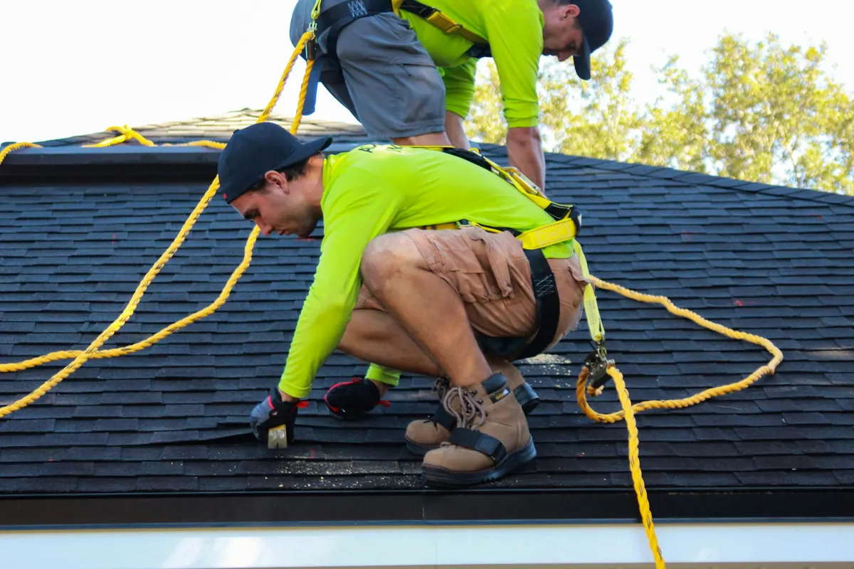 Two roofers in safety harnesses installing shingles on a home